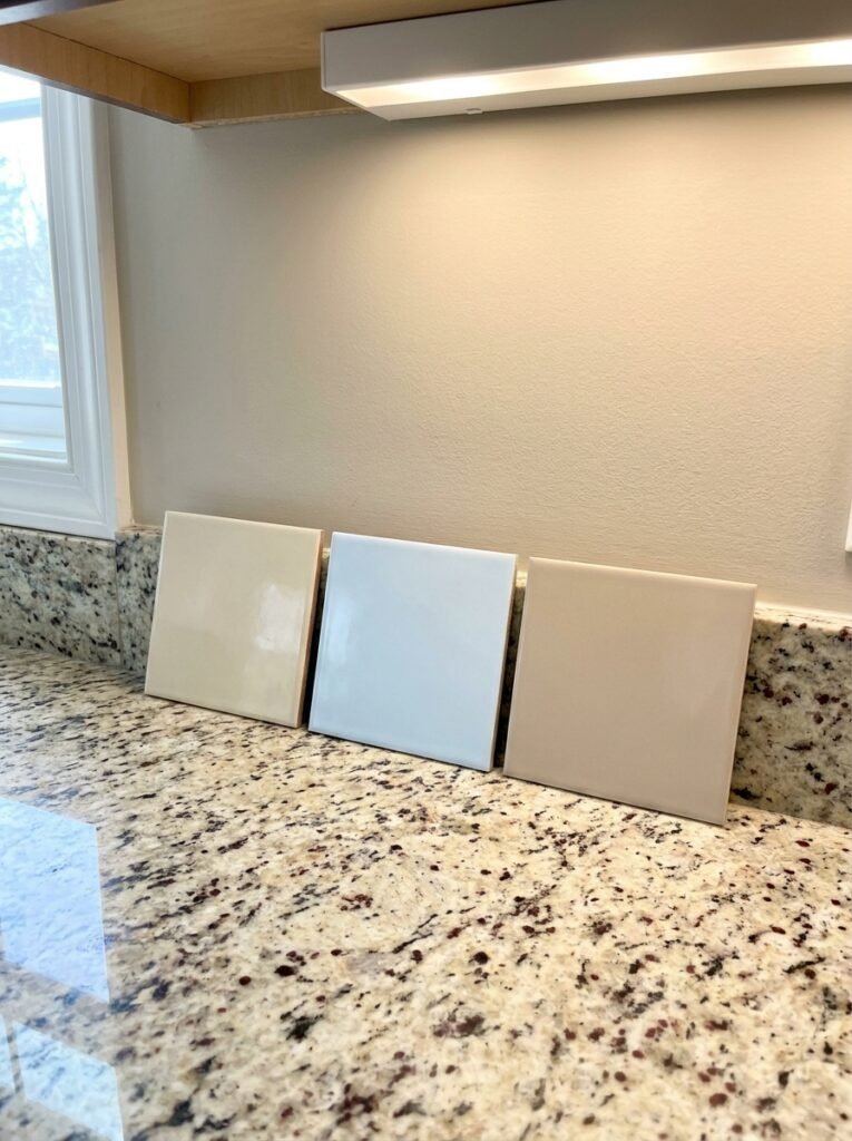 Three tile samples in cream, bright white and soft beige propped against a kitchen wall on a speckled granite counter